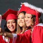 Three students in graduation cap and gown smile for a photo while the one in the middle holds up a phone for a selfie.